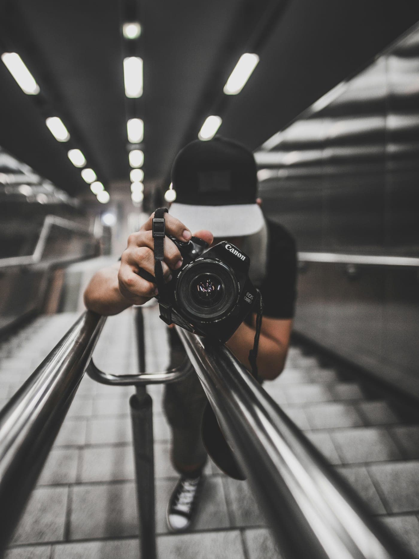 Un photographe dans un couloir de métro, appareil à l’œil, les lignes du plafond convergeant vers lui.